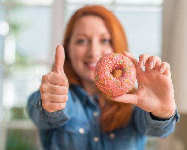 Kızıl saçlı kadın holding çörek, ok işareti yapıyor büyük gülümseme ile mutlu ev, parmaklarıyla, mükemmel işaret parmak