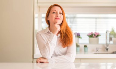 Kızıl saçlı kadın mutfak emin çapraz kollar ve çeneme kaldırdı el ile gülümseme ile kameraya bakıyor. Pozitif düşünce.