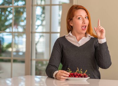 Bir fikir ya da soru işaret parmağı ile mutlu yüz Kızıl saçlı kadın evde kiraz yemek şaşırttı, number one