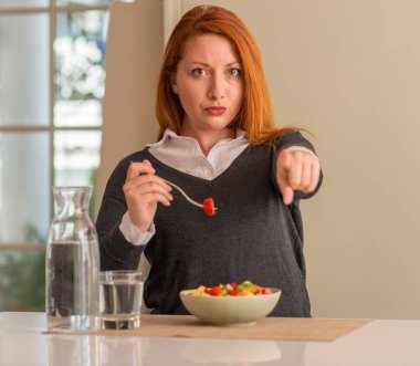 Kızıl saçlı kadın yeme meyve tabağı, kivi ve çilek evde kamera ve size, parmak ile işaret el işareti, pozitif ve kendinden emin bir jest ön