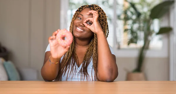 非洲裔美国妇女吃甜甜圈与愉快的面孔微笑做 标志用手在眼睛看通过手指 — 图库照片