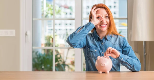 赤毛の女性は 指を通して見る目に手で サインをして笑っている幸せそうな顔で自宅の貯金箱でお金を節約します — ストック写真