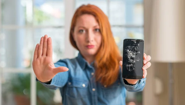 红发女子手持破碎的智能手机用张开的手做停止标志以严肃和自信的表达 防御姿态 — 图库照片