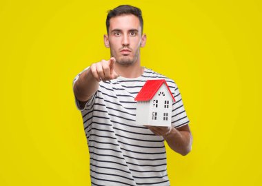 Handsome real estate agent holding a house pointing with finger to the camera and to you, hand sign, positive and confident gesture from the front clipart
