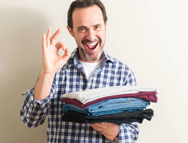 Senior man holding folded laundry clothes doing ok sign with fingers, excellent symbol