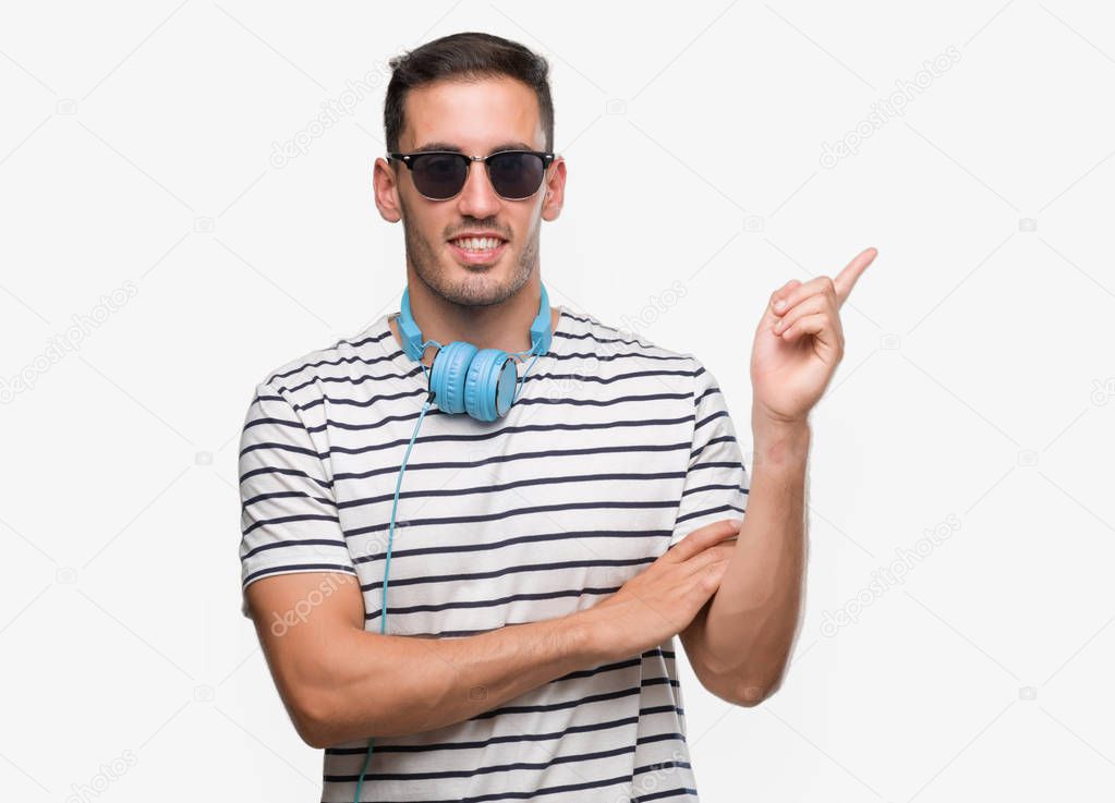 Handsome young man wearing headphones with a big smile on face, pointing with hand and finger to the side looking at the camera.
