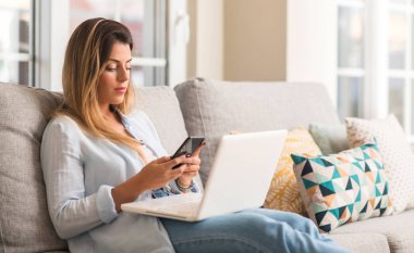 Dizüstü bilgisayar ve telefon kanepede rahat kullanarak gülümseyen genç güzel kadın.