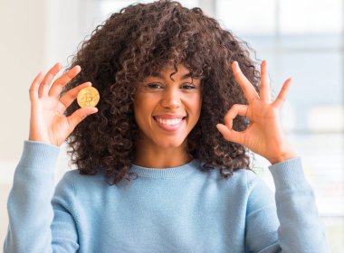 Altın bitcoin cryptocurrency evde tutan Afrikalı-Amerikalı kadın parmakları, mükemmel sembolü olan ok işareti yapıyor