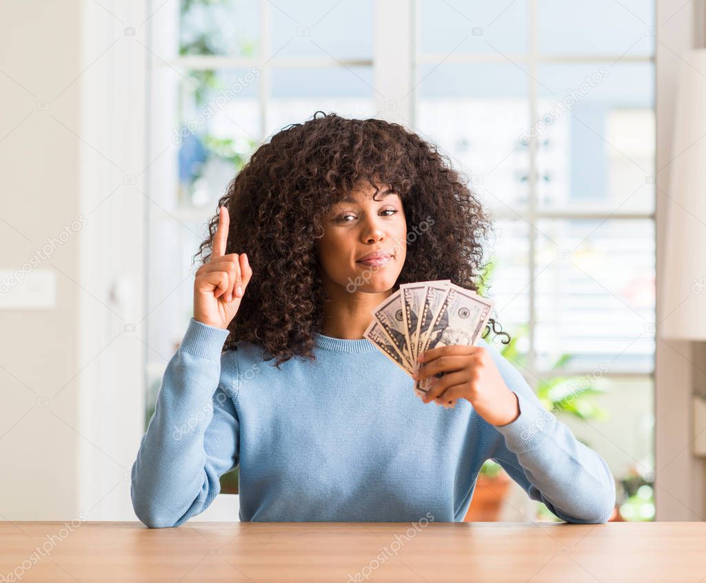 African american woman holding dollar bank notes surprised with an idea or question pointing finger with happy face, number one