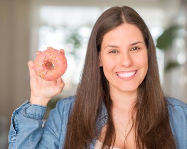 Pembe çörek ile ayakta ve diş gösterilen kendine güvenen bir gülümsemeyle gülümseyerek mutlu bir yüz tutan aç genç kadın