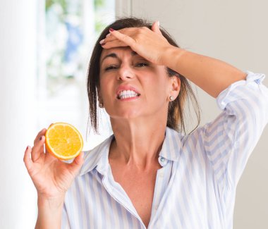 Orta yaşlı elle kafasına, utanç ve sürpriz yüz ile şok vurguladı turuncu meyve tutan kadın öfkeli ve sinirli. Korku ve hata için üzgün.