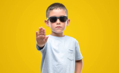 Koyu saçlı küçük çocuk açık eliyle dur işareti ile ciddi ve kendine güvenen ifade, savunma hareketi yaparken güneş gözlüğü takıyor