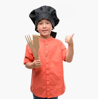 Koyu saçlı küçük çocuk Şef mutlu ok işareti, başparmak ile parmak, mükemmel işareti yapıyor kocaman bir gülümseme ile üniforma