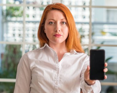 Kızıl saçlı kadın evde Smartphone akıllı yüzünde ciddi düşünme emin bir ifade ile kullanma