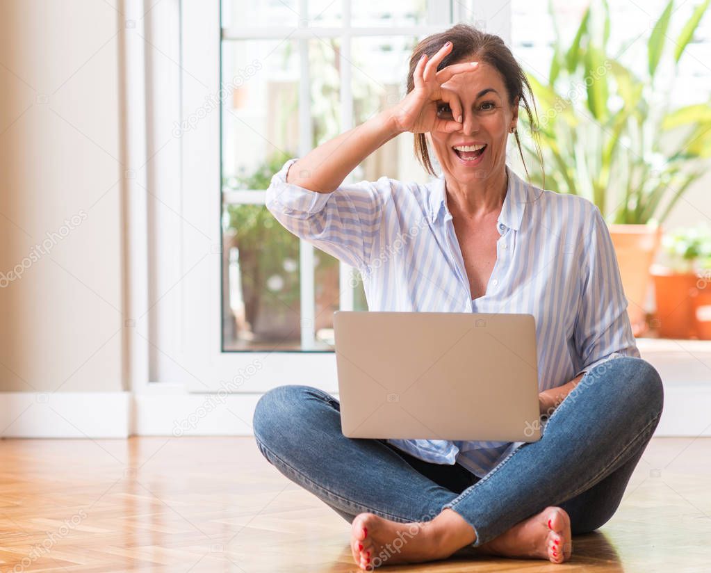 Middle aged woman using laptop at home with happy face smiling doing ok sign with hand on eye looking through fingers