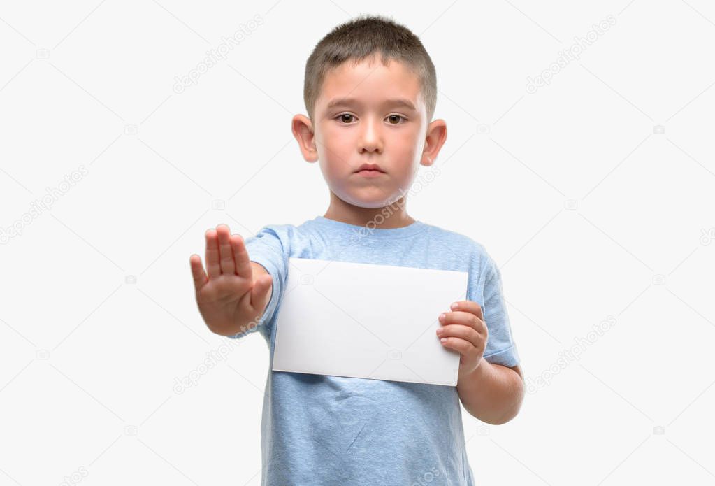Dark haired little child holding a blank card with open hand doing stop sign with serious and confident expression, defense gesture