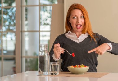 Kızıl saçlı kadın yeme meyve tabağı, kivi ve çilek, çok mutlu el ve parmak ile işaret Anasayfa