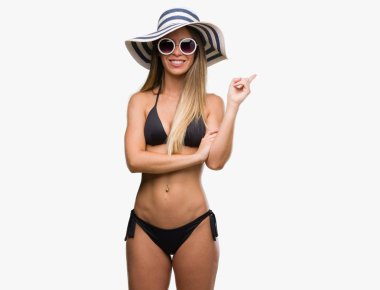 Kameraya bakarak yan el ve parmak ile işaret eden bikini, güneş gözlüğü ve şapka kocaman bir gülümseme ile yüzünde, giyen güzel genç kadın.