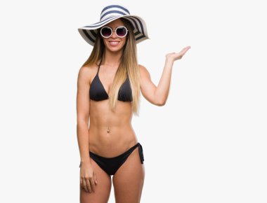 Bikini, güneş gözlüğü ve şapka neşeli sunulması ve kameraya bakarak avucunun ile işaret gülümseyen giyen güzel genç kadın.