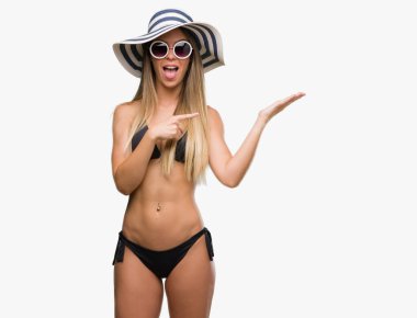 Bikini, güneş gözlüğü ve hayrete ve el ile sunulması ve parmak ile işaret ederken kameraya gülümseyen şapka giyen güzel genç kadın.