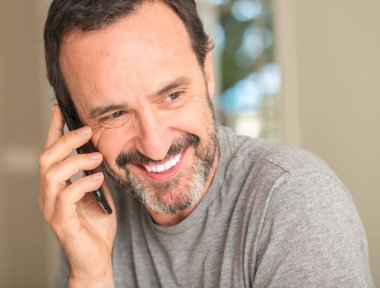 Ayakta ve diş gösterilen kendine güvenen bir gülümsemeyle gülümseyerek mutlu bir yüz ile akıllı telefon kullanan orta yaş adam