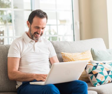 Ayakta ve diş gösterilen kendine güvenen bir gülümsemeyle gülümseyerek mutlu bir yüz ile kanepe dizüstü bilgisayar kullanan orta yaş adam