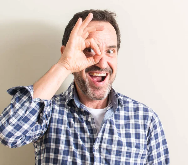 Senior man with happy face smiling doing ok sign with hand on eye looking through fingers