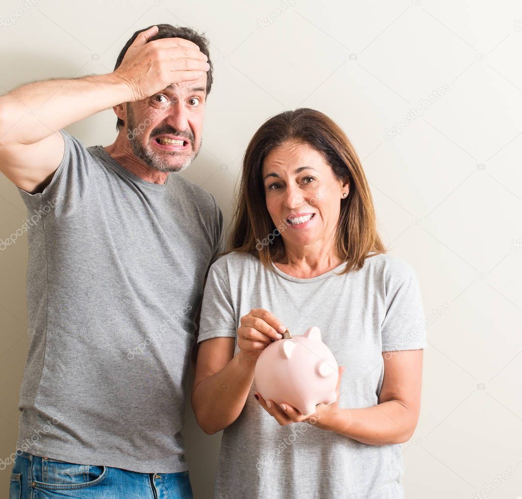 Middle age couple, woman and man, holding piggy bank stressed with hand on head, shocked with shame and surprise face, angry and frustrated. Fear and upset for mistake.