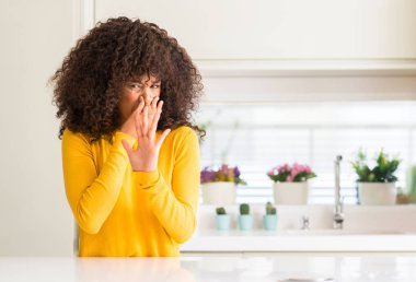 African american woman wearing yellow sweater at kitchen smelling something stinky and disgusting, intolerable smell, holding breath with fingers on nose. Bad smells concept. clipart