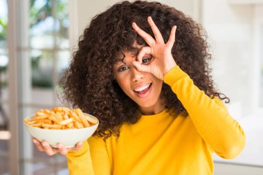 Mutlu yüz el parmaklarının arasından seyir göz üzerinde ok işaretiyle yapıyor gülümseyen bir tabak patates cipsi evde ile tutan Afrikalı-Amerikalı kadın