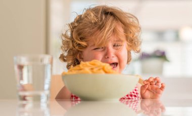 Beautiful blond child eating spaghetti with hands crying with tantrum at home. clipart