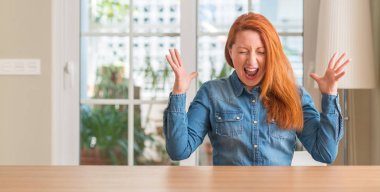 Redhead woman at home celebrating mad and crazy for success with arms raised and closed eyes screaming excited. Winner concept clipart