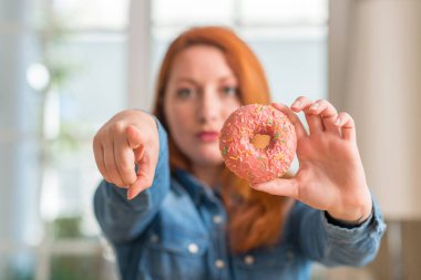 Kızıl saçlı kadın holding çörek evde işareti, pozitif ve kendinden emin bir jest ön el kamera ve size, parmak ile işaret