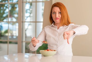 Kızıl saçlı kadın yeme tahıllar ile ahududu evde kızgın yüzü ile işaret gösteren antipati başparmak aşağı, ret kavramı ile negatif.