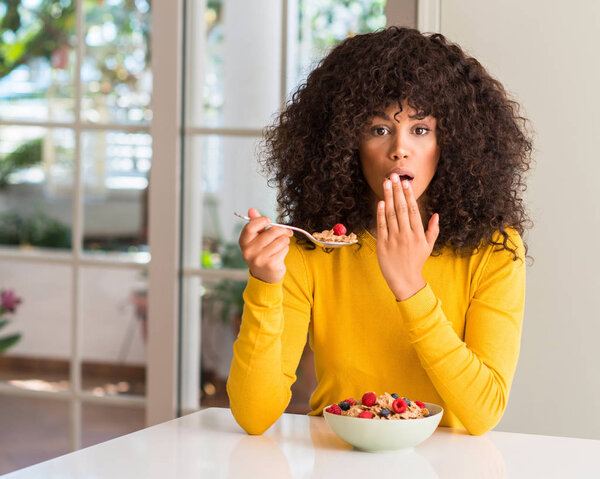 African american woman eating cereals, raspberries and blueberries cover mouth with hand shocked with shame for mistake, expression of fear, scared in silence, secret concept