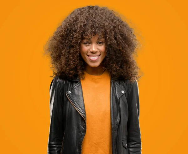 Afrikalı-Amerikalı kadın ayakta ve diş gösterilen kendine güvenen bir gülümsemeyle gülümseyerek mutlu bir yüz ile deri ceket