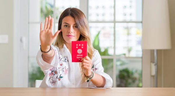Young woman at home holding a passport of Japan with open hand doing stop sign with serious and confident expression, defense gesture