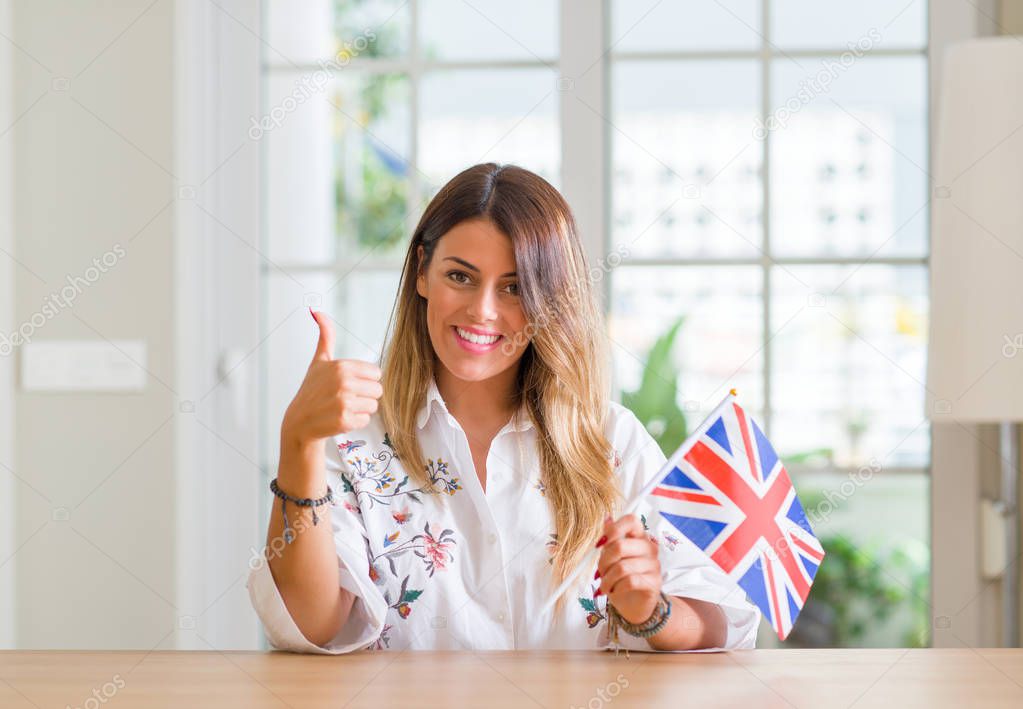 Young woman at home holding flag of United Kingdom happy with big smile doing ok sign, thumb up with fingers, excellent sign