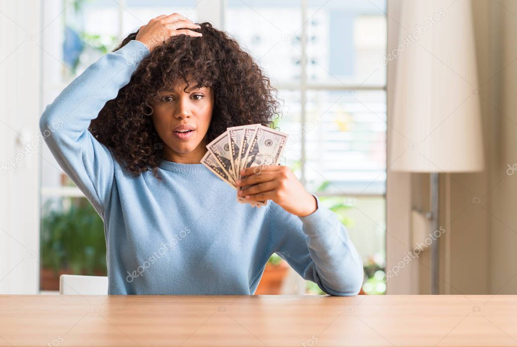 African american woman holding dollar bank notes stressed with hand on head, shocked with shame and surprise face, angry and frustrated. Fear and upset for mistake.