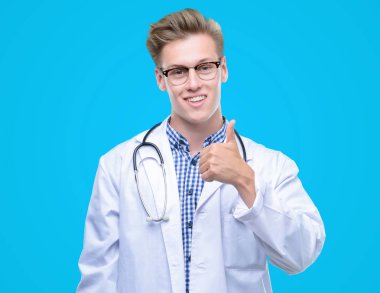 Genç yakışıklı sarışın doktor mutlu ok işareti, başparmak ile parmak, mükemmel işareti yapıyor kocaman bir gülümseme ile