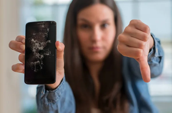 Brunette young woman holding broken smartphone with angry face, negative sign showing dislike with thumbs down, rejection concept