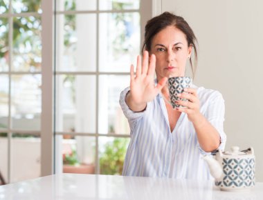 Dur işareti ile ciddi ve kendine güvenen ifade, savunma hareketi yaparken açık el ile bir fincan çay ya da kahve içme orta yaşlı kadın