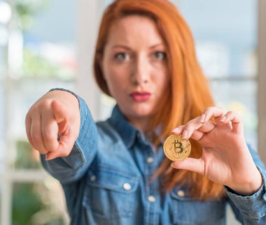 Bitcoin cryptocurrency evde tutan Kızıl saçlı kadın kamera ve size, el işareti, pozitif ve kendinden emin bir jest ön parmak ile işaret