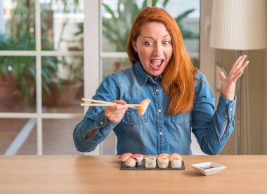 Kocaman bir gülümseme ile çığlık zaferini kutluyor chopsticks çok mutlu ve heyecanlı, kazanan ifade kullanarak suşi yemek ve ellerini kaldırdı Kızıl saçlı kadın