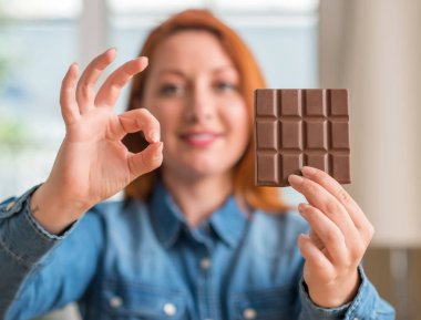 Parmak, mükemmel sembolü ile ok işareti yapıyor evde çikolata bar tutan Kızıl saçlı kadın