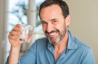Orta yaş adam ayakta ve diş gösterilen kendine güvenen bir gülümsemeyle gülümseyerek mutlu bir yüz ile bir bardak su içme