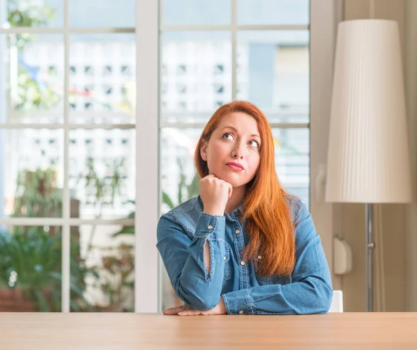 Redhead woman at home with hand on chin thinking about question, pensive expression. Smiling with thoughtful face. Doubt concept.