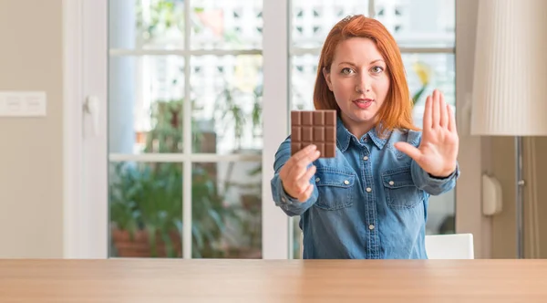 Redhead woman holding chocolate bar at home with open hand doing stop sign with serious and confident expression, defense gesture
