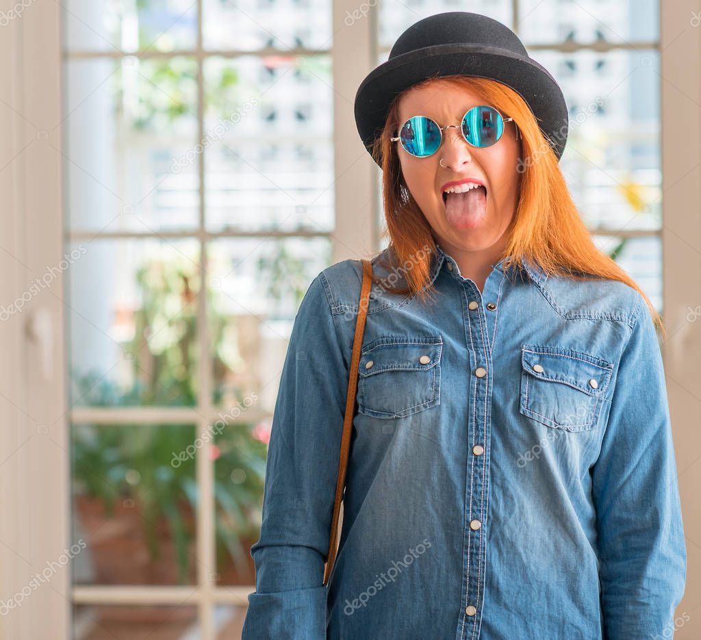 Stylish redhead woman wearing bowler hat and sunglasses sticking tongue out happy with funny expression. Emotion concept.