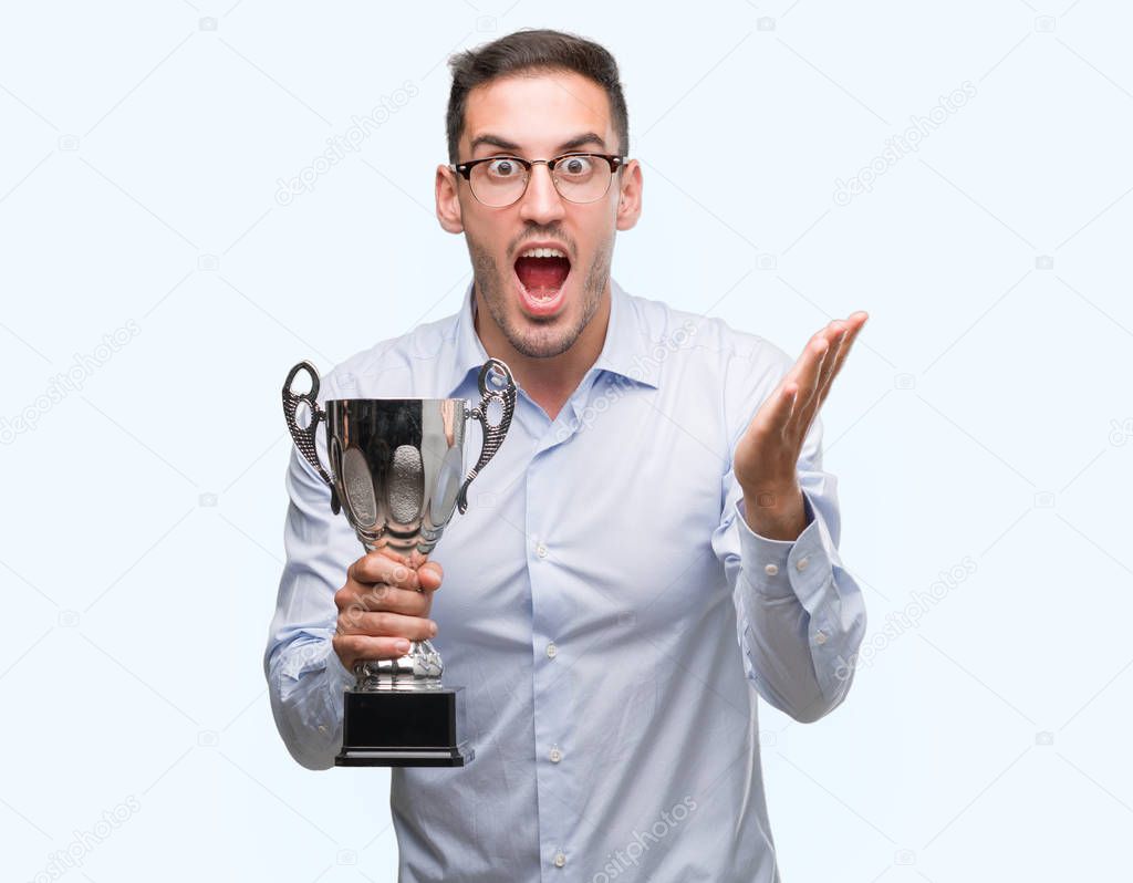 Handsome young man holding trophy very happy and excited, winner expression celebrating victory screaming with big smile and raised hands